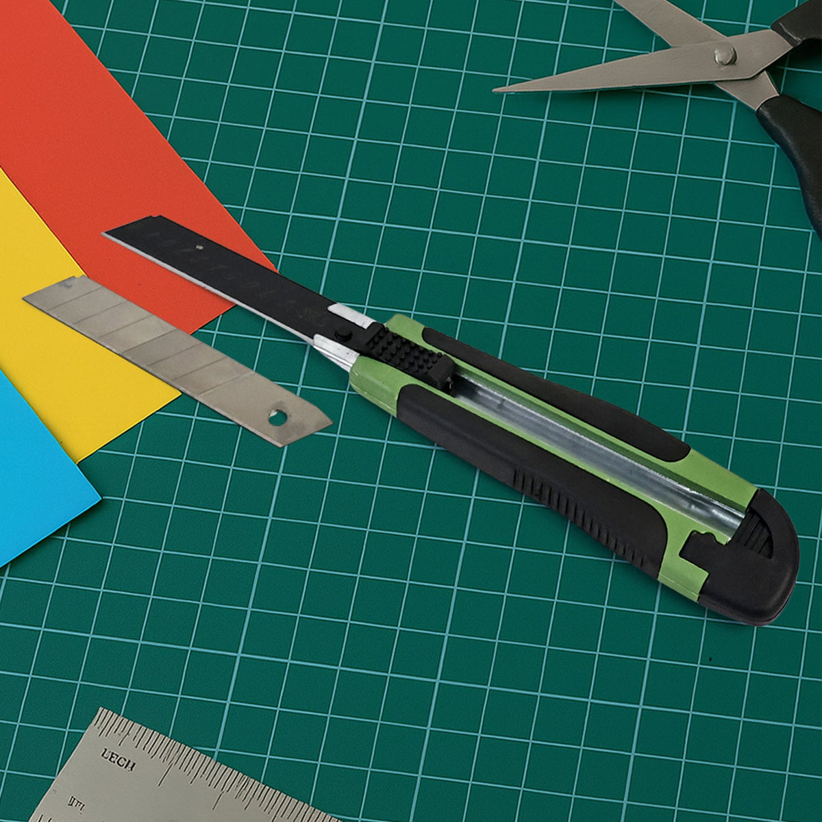 A green and black utility knife with a loose blade and colorful paper on a green cutting mat.