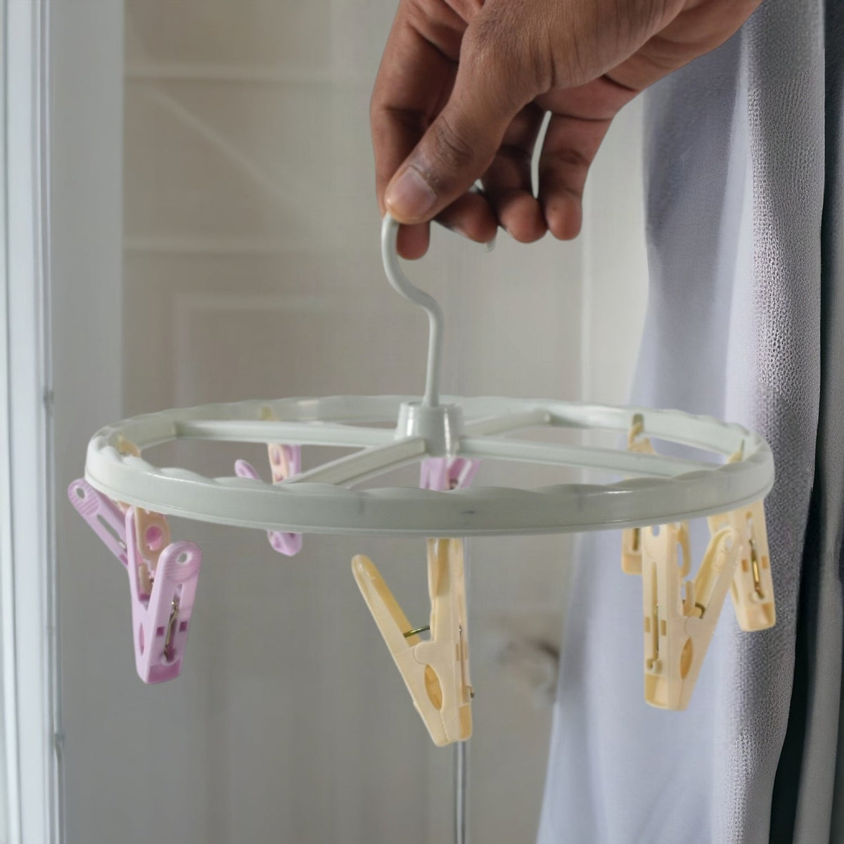A hand holds a light blue circular drying rack with purple and yellow clips indoors.