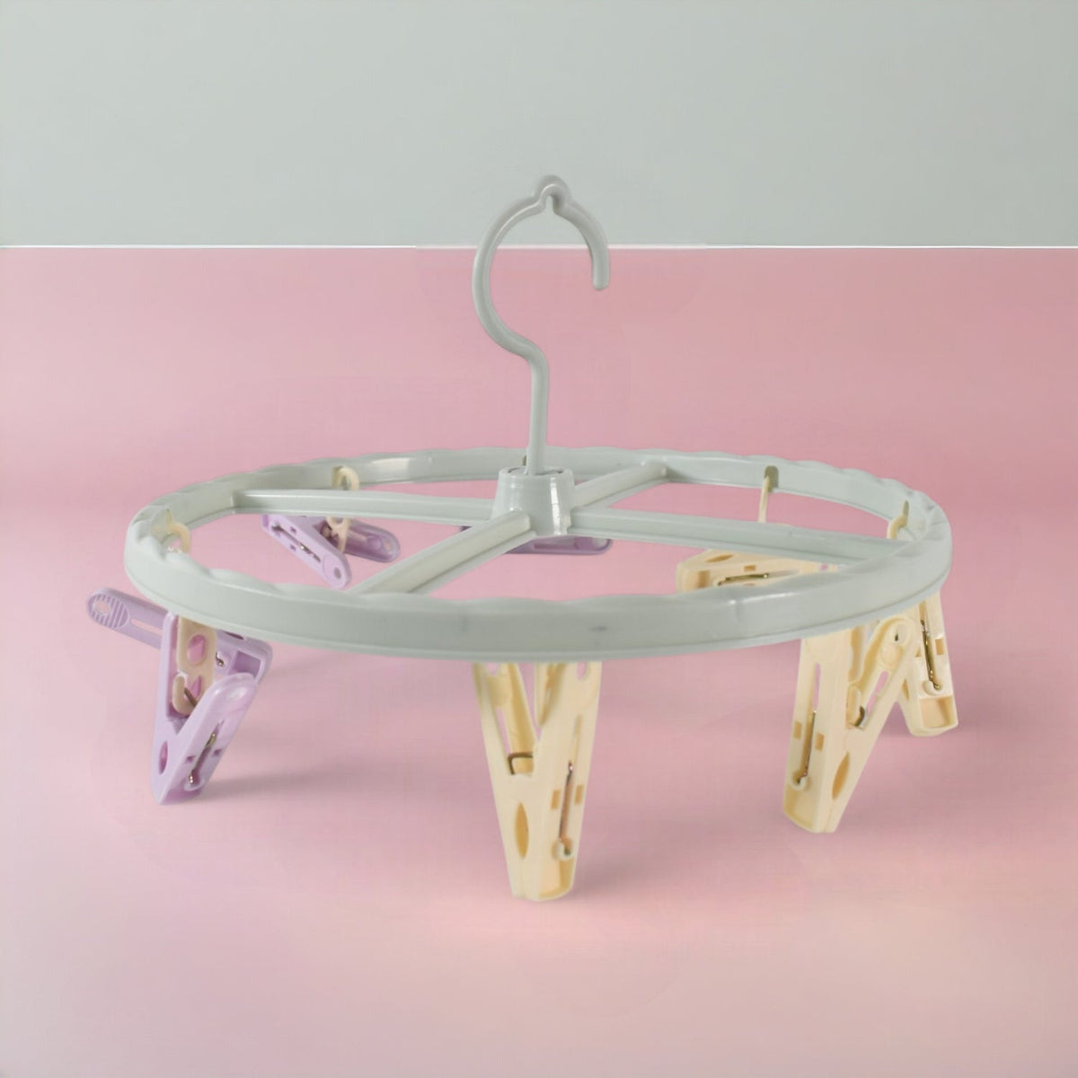 A light blue circular drying rack with purple and yellow clips is centered on a pink and grey background.