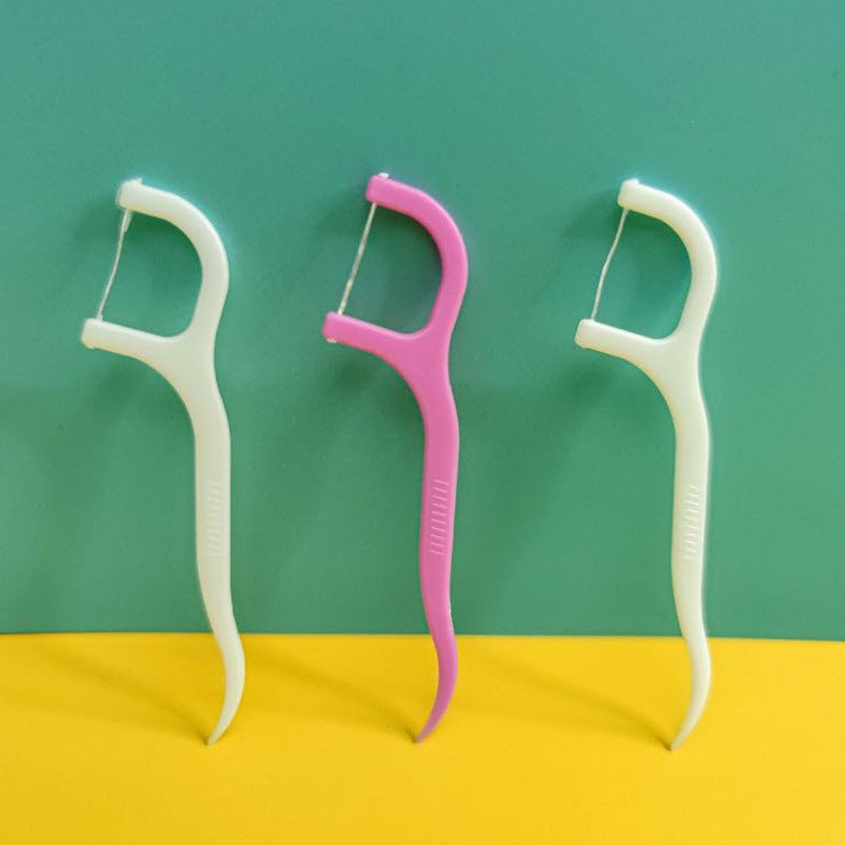Three dental floss picks, two white and one pink, standing upright against a two-toned teal and yellow background.