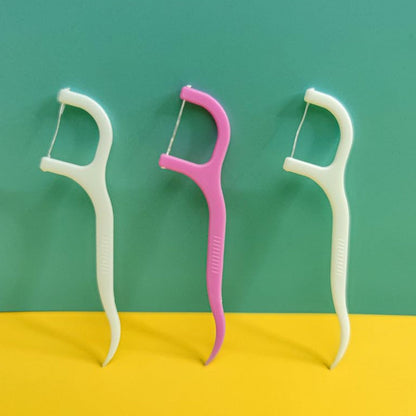 Three dental floss picks, two white and one pink, standing upright against a two-toned teal and yellow background.