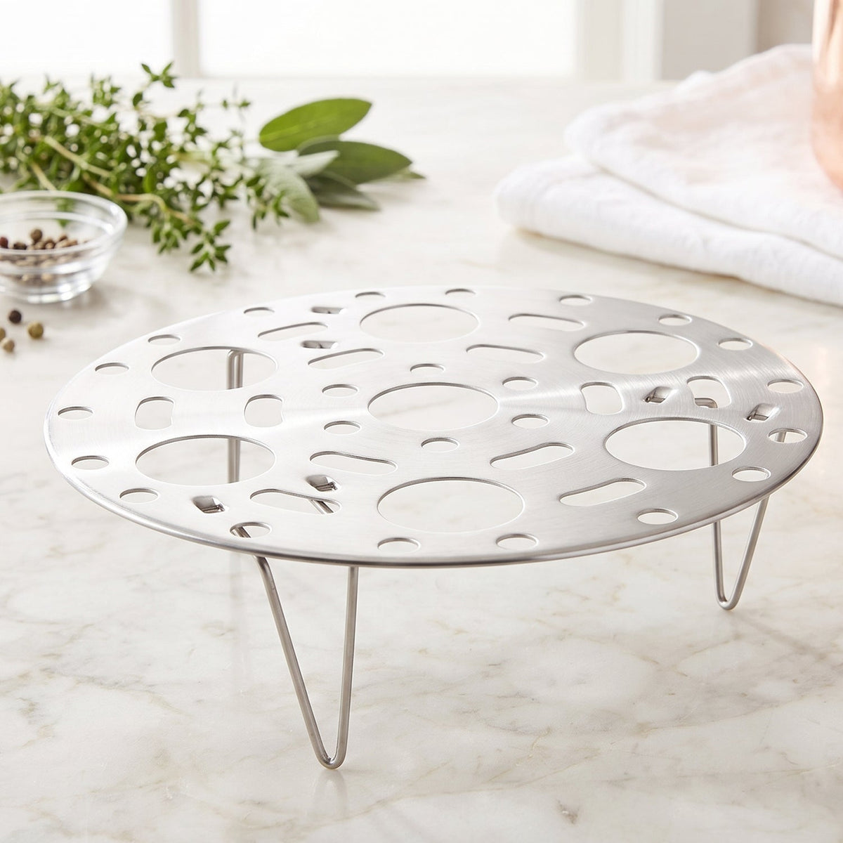 A polished metal egg steamer rack with three legs sits on a bright marble countertop, surrounded by green herbs.