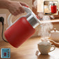 A hand pours steaming hot water from a red ribbed electric kettle into a white teacup with a tea bag. The kettle has a white