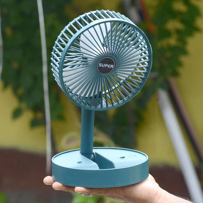 A hand holds a dark green telescopic foldable mini USB desk fan, extended, against an outdoor background.