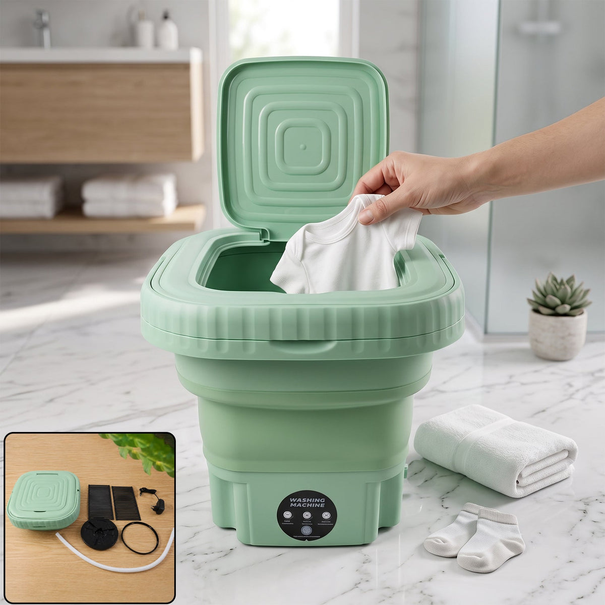 A hand places a white baby onesie into a green portable washing machine with its lid open, next to folded towels and baby