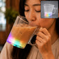A person sips an iced coffee from a clear heart-shaped mug with a rainbow-lit base. A smaller inset shows the mug empty.