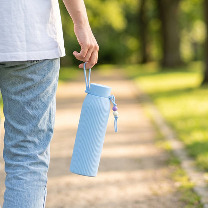 A person walks outdoors, holding a light blue ribbed glass water bottle with a beaded strap by their side.