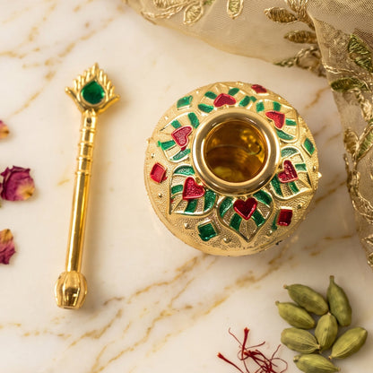 The gold decorative pot and spoon, both with red and green enamel, are displayed on a white marble surface with spices.