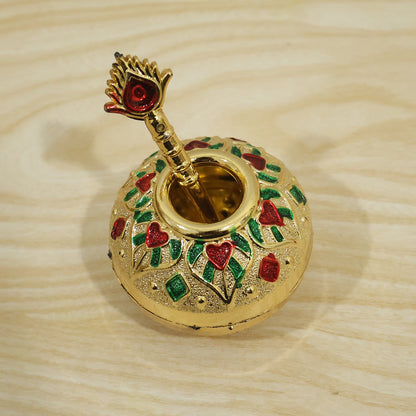 A gold decorative pot and spoon, with red and green enamel, are shown on a light wooden surface.