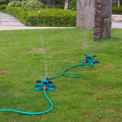Two blue and black sprinklers connected by a green hose, watering a lush green lawn in a park setting.