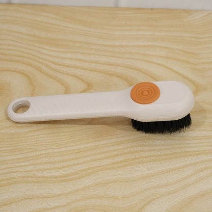 A white scrubbing brush with black bristles and an orange button lies on a light wooden surface.