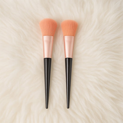 Two large, soft, peach-colored makeup brushes with black handles lie side by side on a white fluffy texture.