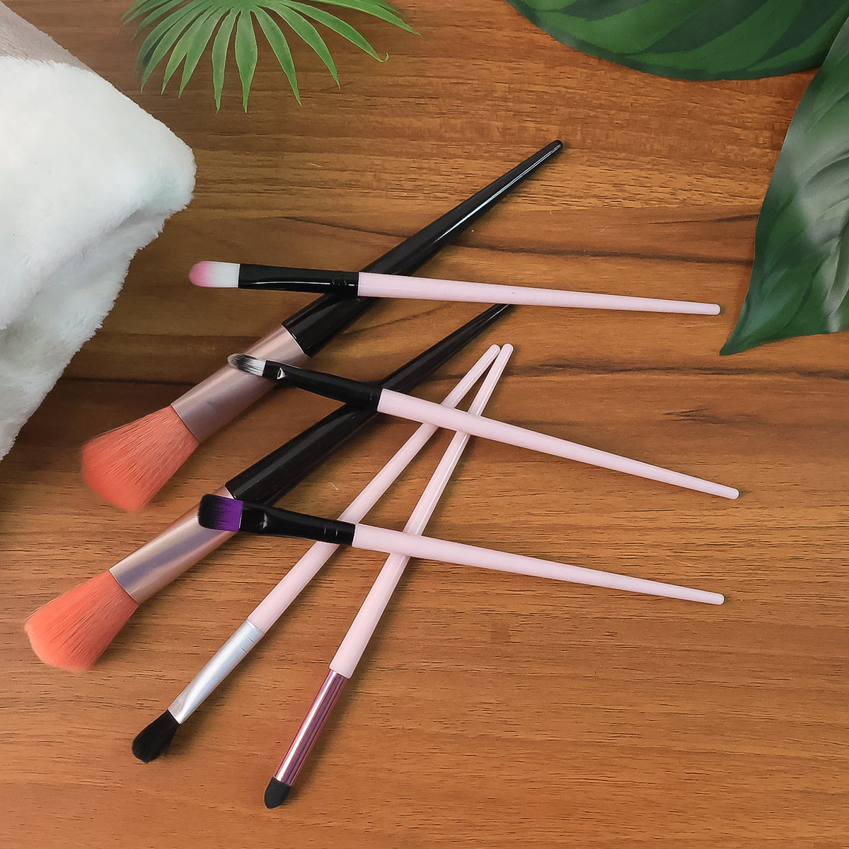 Seven makeup brushes with pink and black handles are scattered on a wooden surface with green leaves.