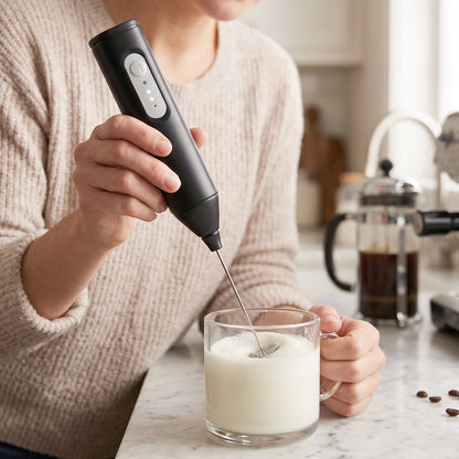 A person in a cream sweater uses a black electric milk frother to create foam in a clear glass of milk on a kitchen counter.