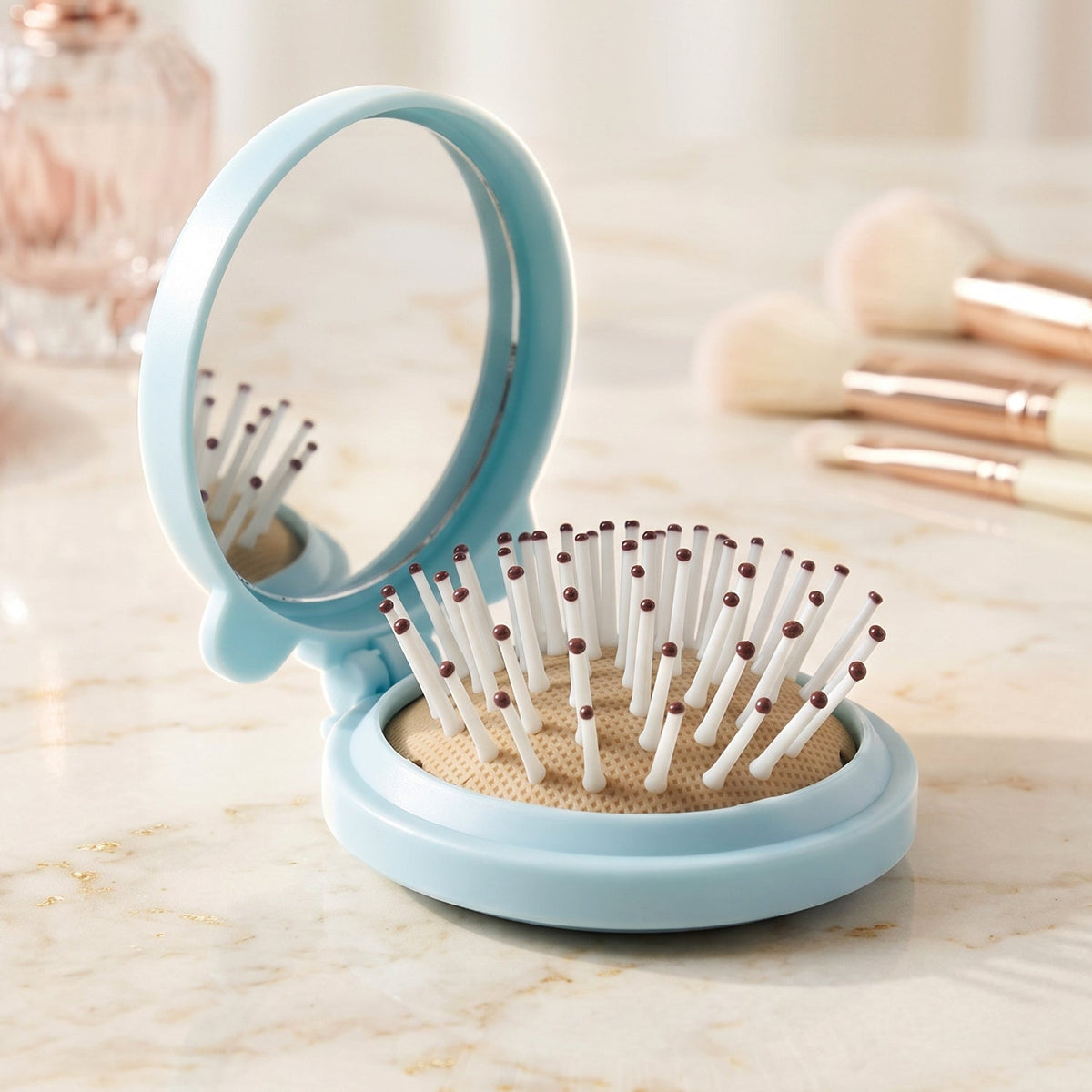 A light blue folding hairbrush with a mirror stands open on a marble surface, with makeup brushes in the soft background.