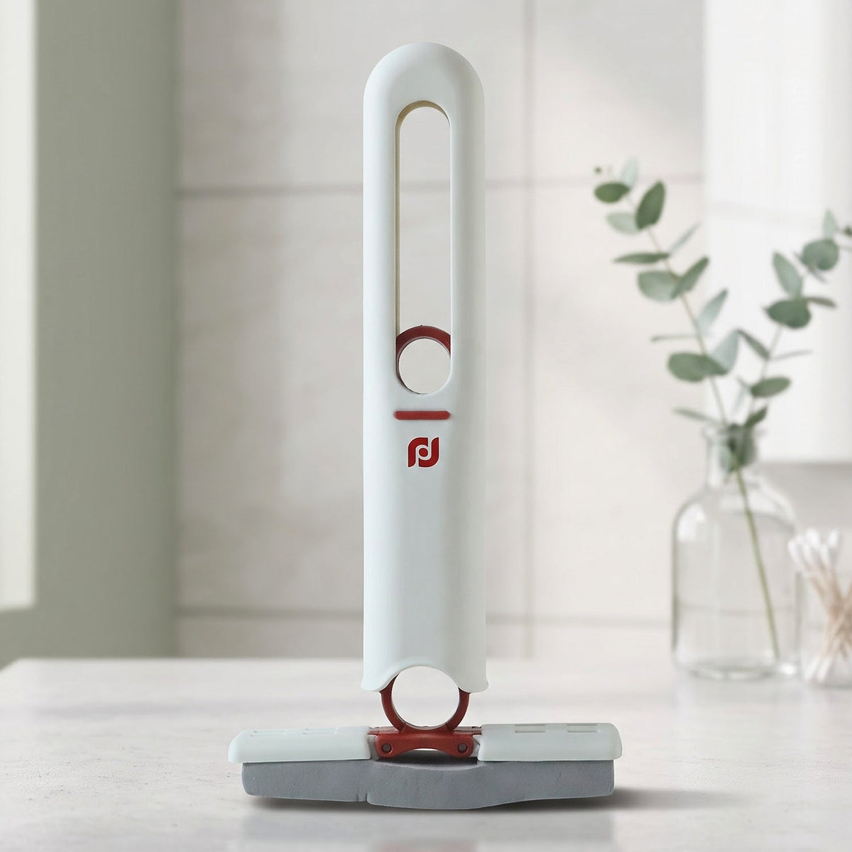 A white and red mini mop with a grey sponge head stands upright on a white countertop with a plant in the background.