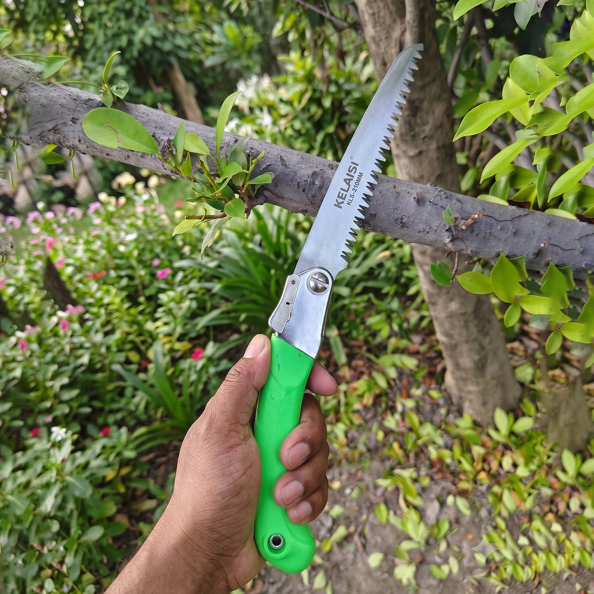 A hand holds the green handled folding saw, extending it towards a tree branch in a garden setting.