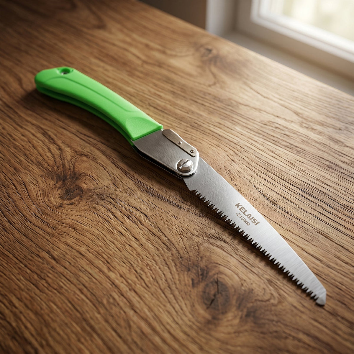 A green handled folding saw with a silver blade is displayed on a polished wooden surface near a window.