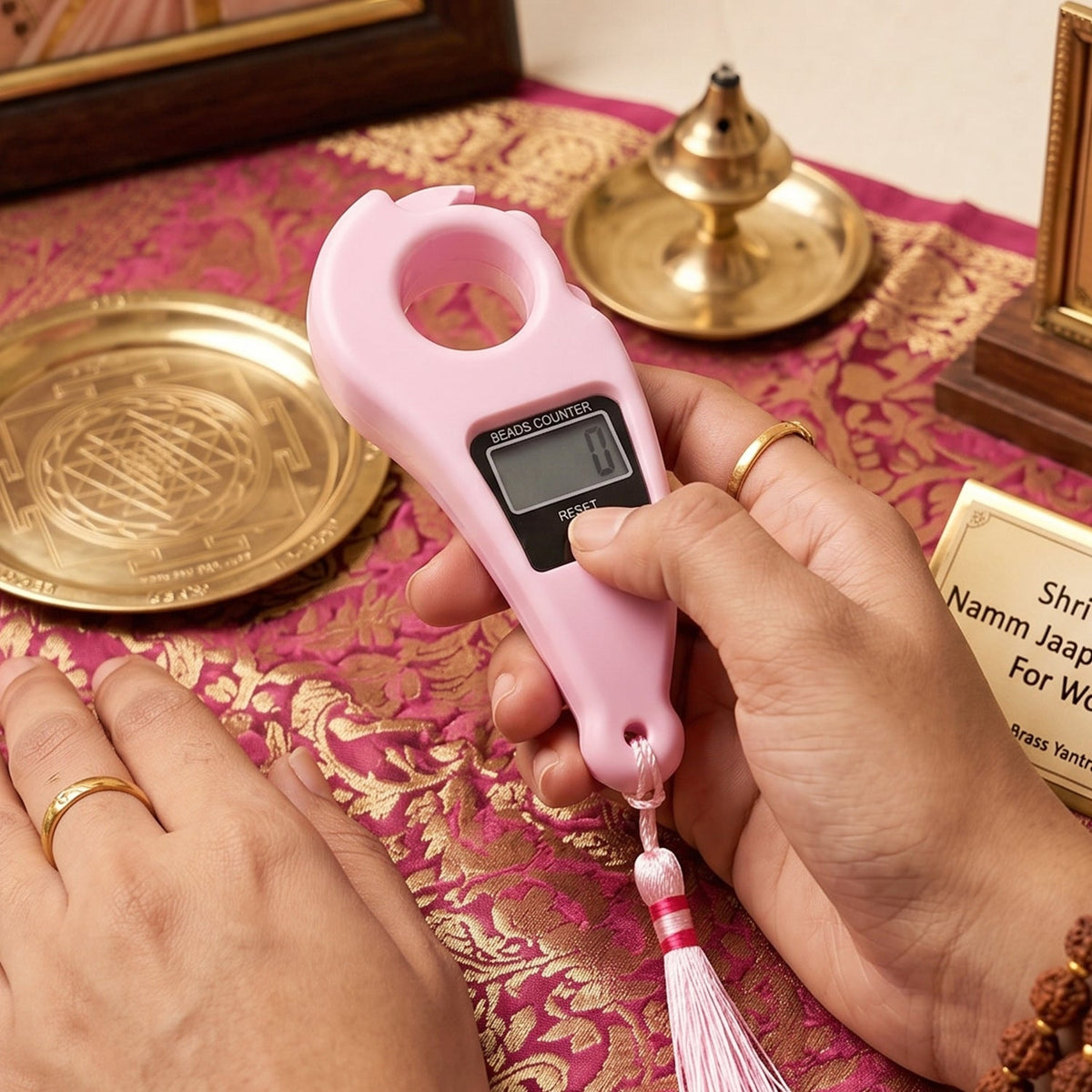 Hands operating the pink digital beads counter, surrounded by traditional brass religious items on a patterned fabric.