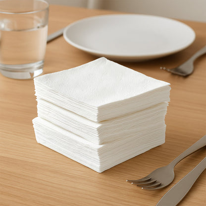 A stack of white paper napkins on a wooden table with a glass of water and a plate.