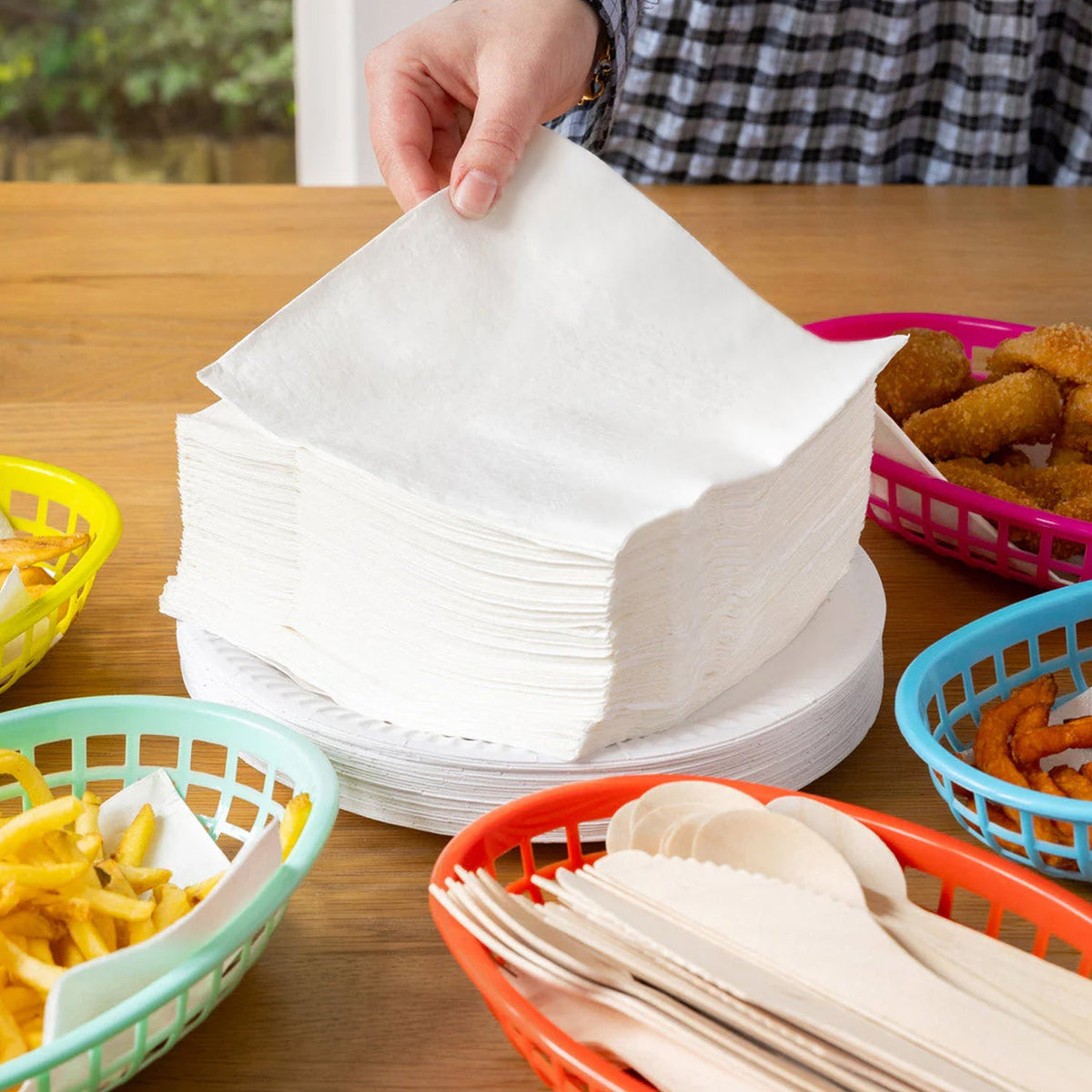 A hand reaches for a white paper napkin from a large stack, surrounded by colorful baskets of food.