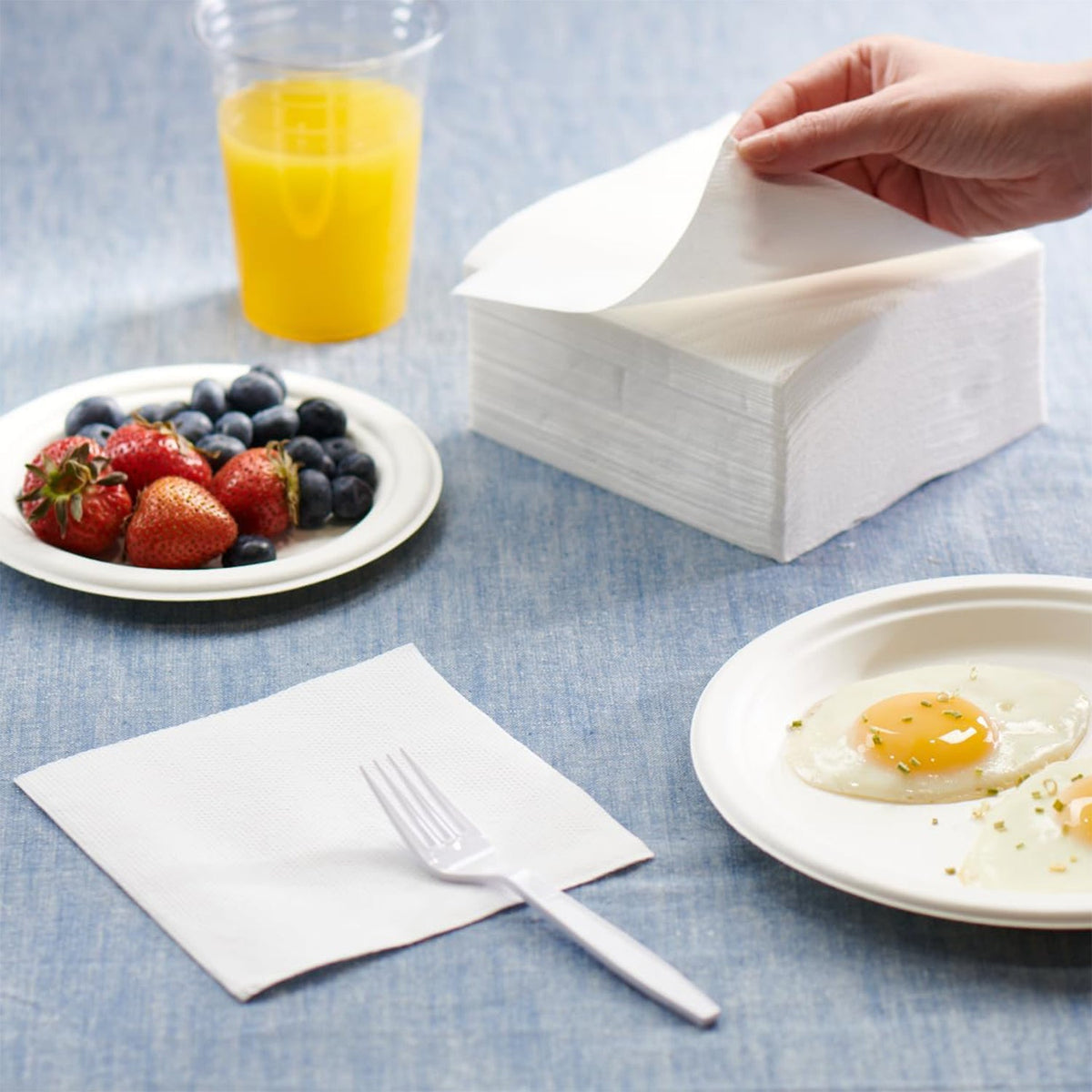 A hand pulls a white paper napkin from a stack, with fruit, eggs, and orange juice nearby.
