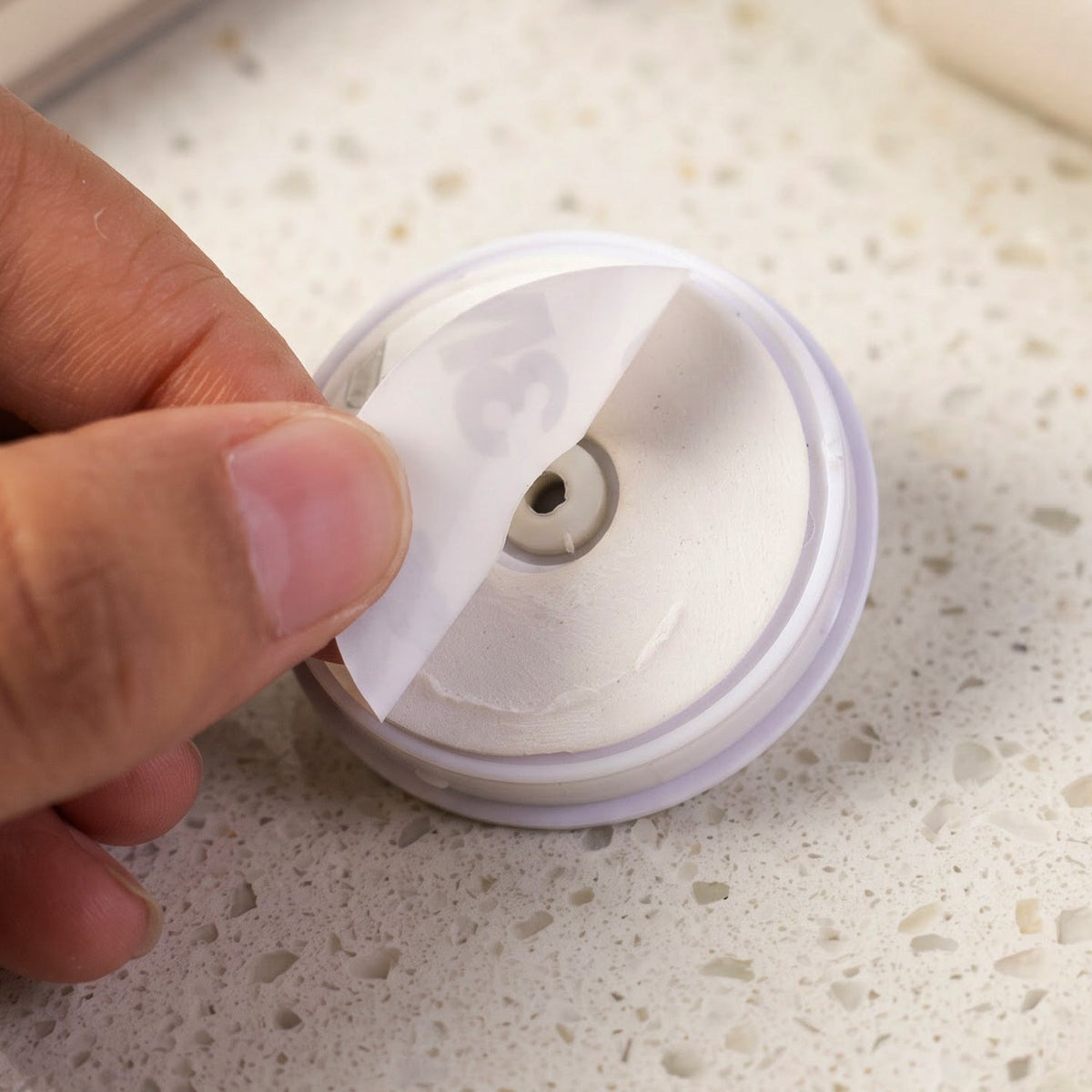 A hand peels off the protective backing from the adhesive side of a white pop socket base.