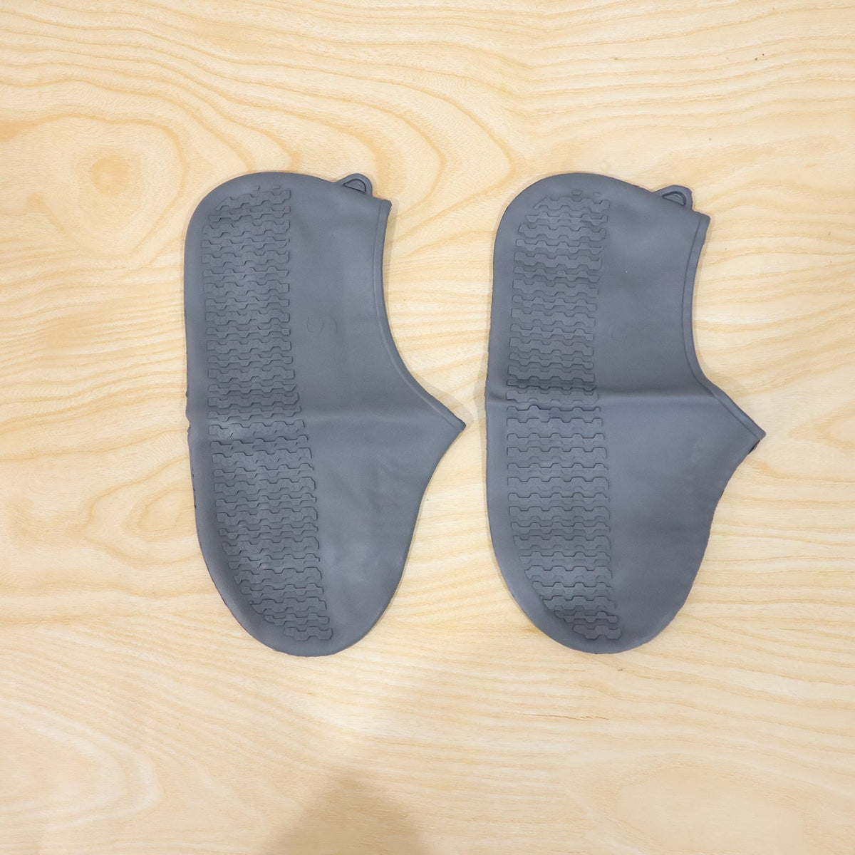 Two dark grey silicone shoe covers with textured soles are laid flat on a light wooden surface.