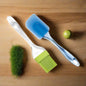 A blue silicone spatula and a green silicone pastry brush lie on a light wooden surface with a green moss-like object and a