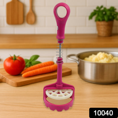 A pink and white plastic potato masher stands upright on a wooden counter with vegetables in the background.