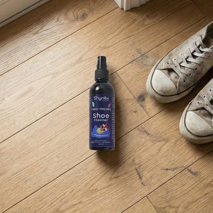 A Shynex shoe cleaner bottle stands on a wooden floor next to a pair of dirty grey canvas shoes.