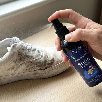 A hand sprays Shynex shoe cleaner onto a heavily soiled white canvas shoe near a window.
