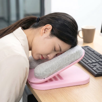 A woman with dark hair sleeps on a pink desktop nap pillow with a gray cushion at an office desk.