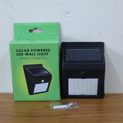 Black solar-powered LED wall light next to its green retail box and mounting screws on a wooden surface.