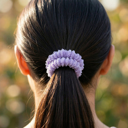A close-up of a child's dark hair tied in a ponytail with a purple fluffy elastic hair band against a blurred outdoor