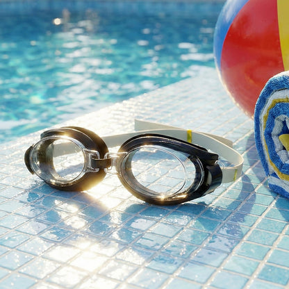 Black and clear swimming goggles with a beige strap on blue pool tiles next to a beach ball and towel.