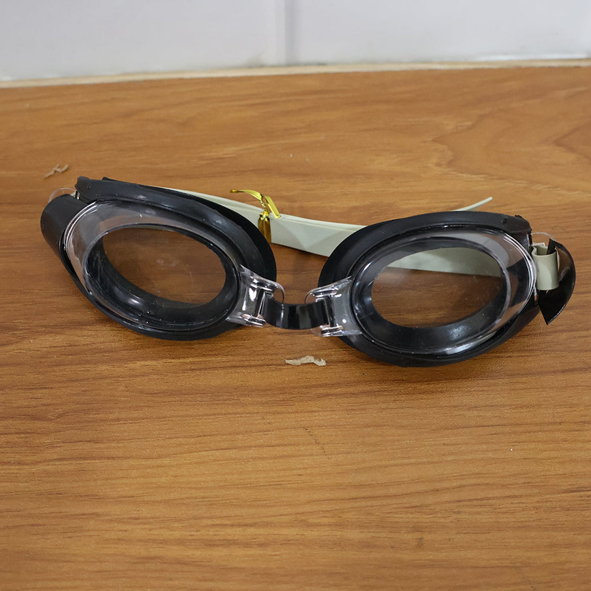 Black and clear swimming goggles with a beige strap resting on a light brown wooden surface.