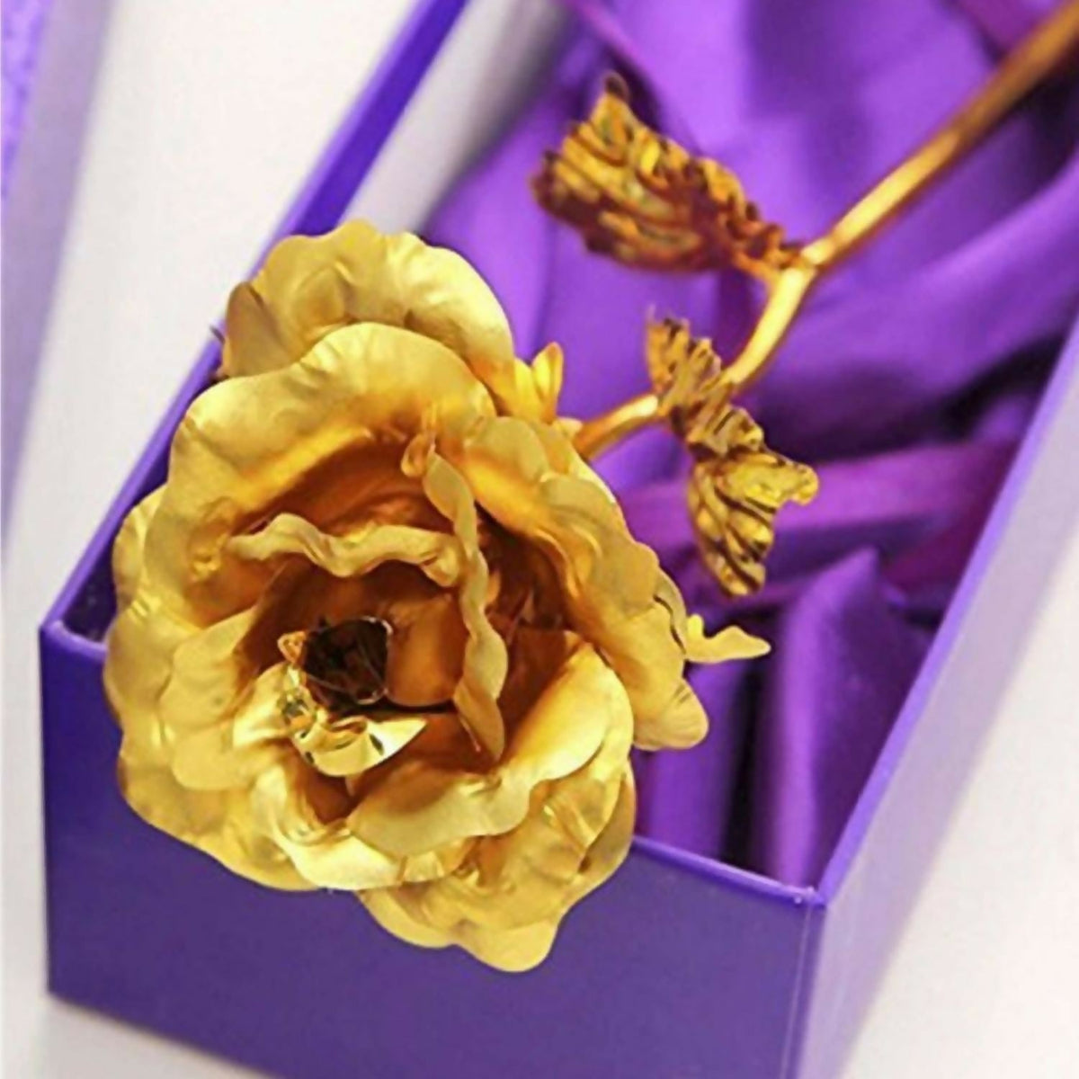 A close-up of a gold-plated rose nestled inside an open purple gift box with a satin lining.