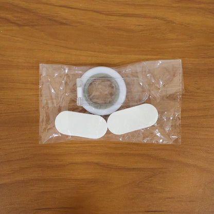 A clear plastic package containing a white and gray toilet seat lifter and two white oval adhesive pads on a wooden surface.