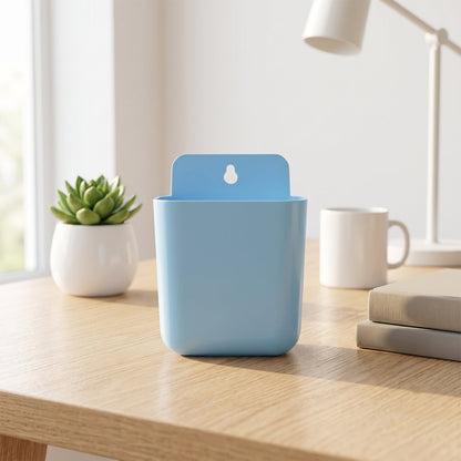 A light blue wall-mounted plastic storage pocket sits on a wooden desk with a plant, mug, and books in a bright room.