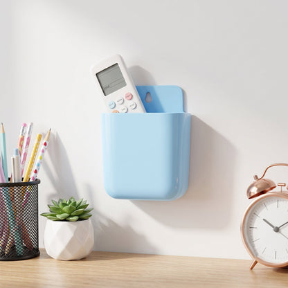A light blue wall-mounted plastic storage pocket holds a white remote control above a desk with a plant, pencils, and a rose