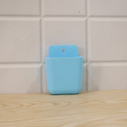 A light blue wall-mounted plastic storage pocket is shown against a white tiled wall and a wooden surface.