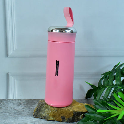 A pink vacuum flask with a silver lid and strap sits on a rock with green leaves and a white wall in the background.