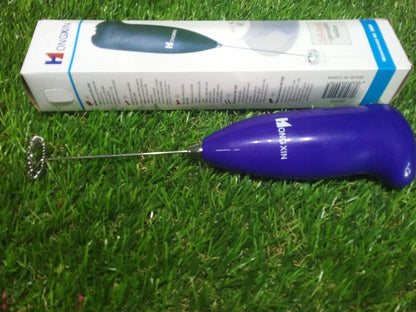 A purple frother and its white and blue packaging box are displayed on a vibrant green grass background.
