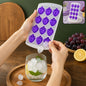 Hands removing a clear fruit-shaped ice cube from a purple silicone tray into a glass with ice and lemon.