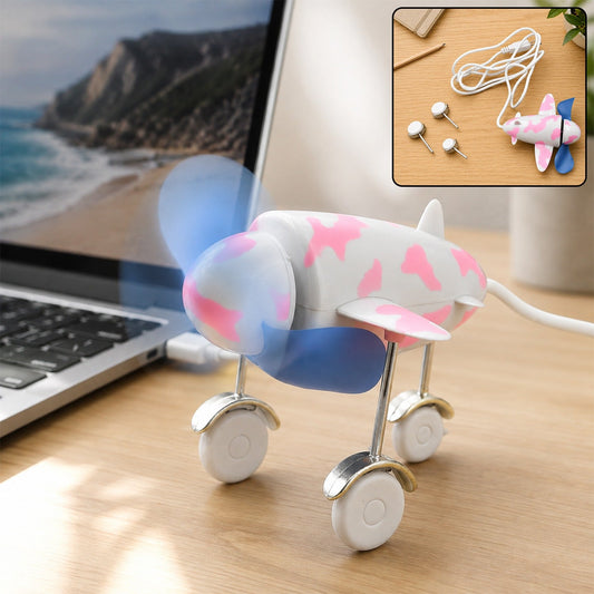 A pink and white airplane-shaped USB fan with blue propellers is plugged into a laptop on a wooden desk.