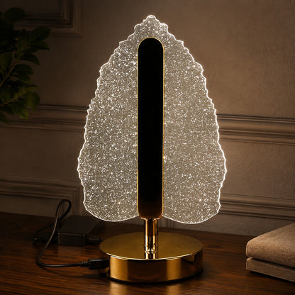 Illuminated crystal leaf lamp with a gold base and stem, plugged in on a dark wooden surface.