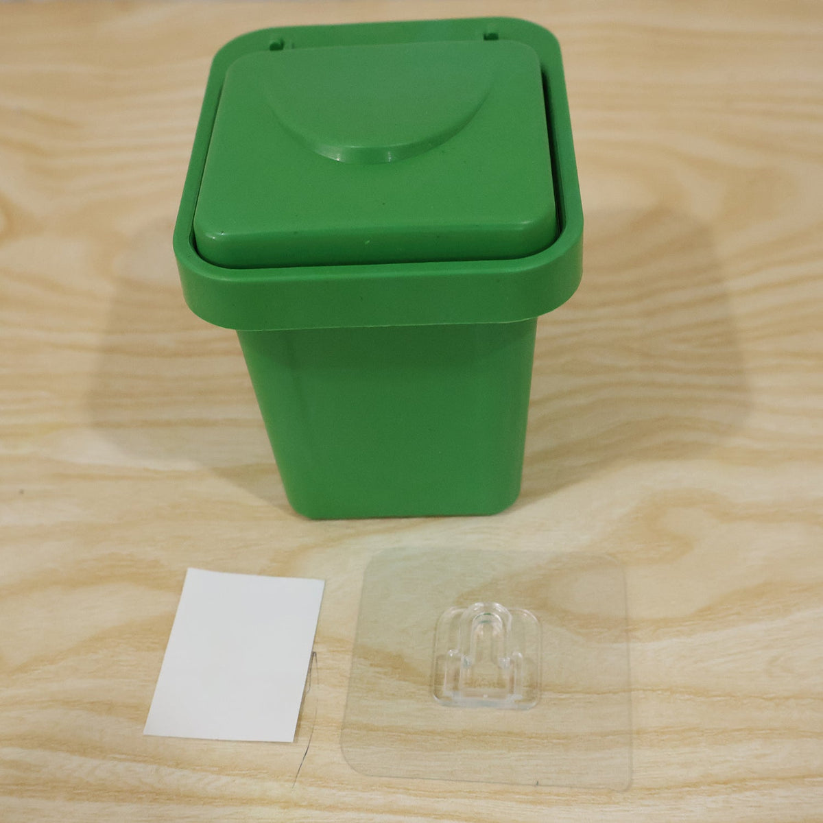 A closed green mini trash can ashtray on a wooden surface with an adhesive wall mount and a small white sticker.