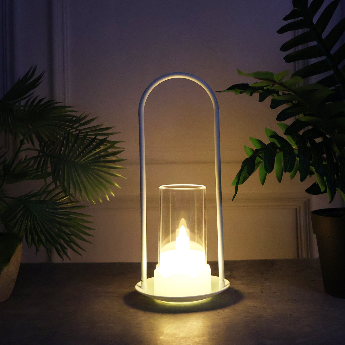 A single white LED wind lantern with a glowing flameless candle, placed on a dark surface with green plants in the