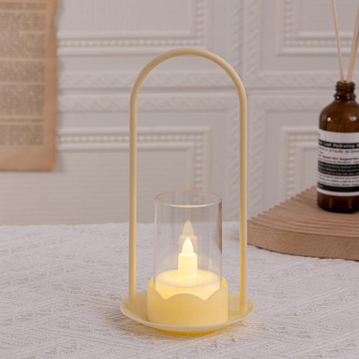 A single light yellow LED wind lantern with a glowing flameless candle, on a white textured surface with a brown bottle in